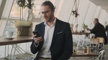 Yakışıklı, genç, sakallı bir iş adamı, kapalı bir kafede kahve içerken akıllı telefonunu kullanıyor.