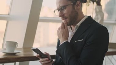 Ciddi bir genç sakallı iş adamı kapalı bir kafede kahve içerken akıllı telefonunu kullanıyor.