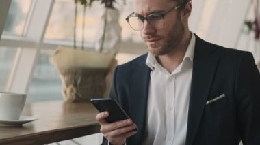 Sakallı sakallı genç bir işadamı, kapalı bir kafede kahve içerken akıllı telefonunu kullanıyor.