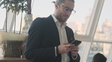 Memnun bir işadamı akıllı telefonunu kapalı bir kafede beklerken ve beklerken kullanıyor.