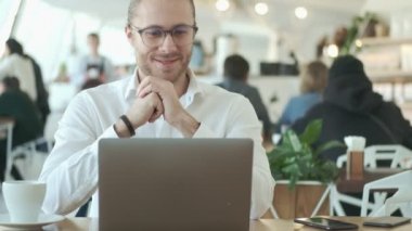 Yakışıklı, pozitif, gözlüklü genç bir işadamı bilgisayarını kullanıyor ve kapalı bir kafede kahve içerken video bağlantısıyla konuşuyor.