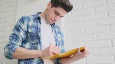Odaklanmış bir adam, elinde kağıt bir klasör tutarak evde dikilirken notlar alıyor.