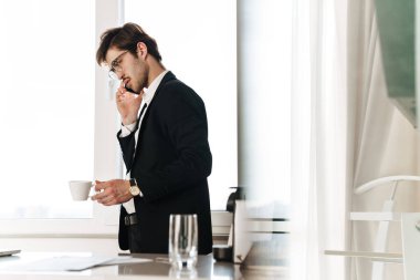 Modern mutfakta kahve içerken cep telefonu kullanan gözlüklü genç işadamının fotoğrafı.