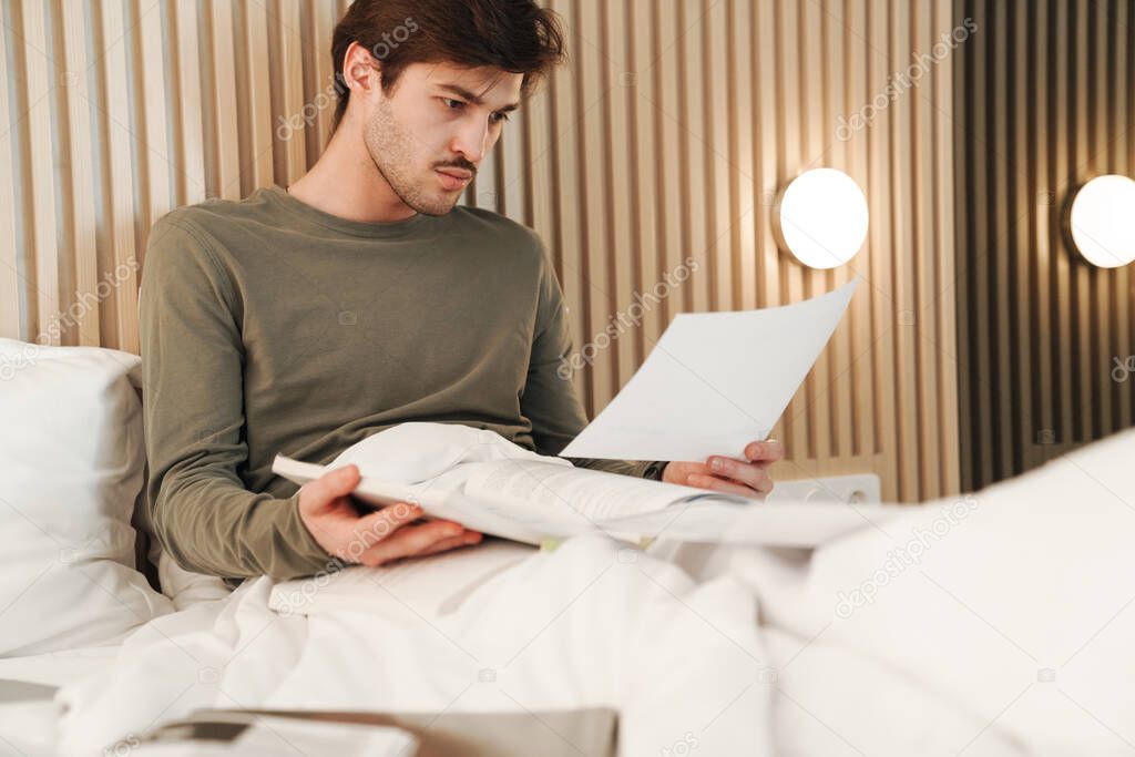Photo of caucasian focused man with mustache reading documents while working on bed in apartment