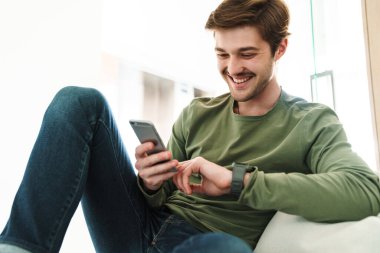 Bıyıklı, gülümseyen ve akıllı telefon kullanan neşeli genç adamın fotoğrafı.