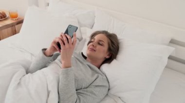Oldukça hoş genç bir kadın, beyaz yatakta yatarken akıllı telefonundan güzel bir şey izliyor.