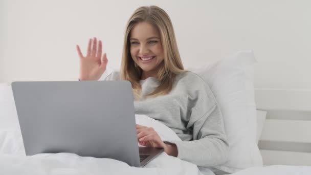 彼女のラップトップコンピュータを使用してビデオと話しています彼女の白いベッドで自宅で朝 — ストック動画