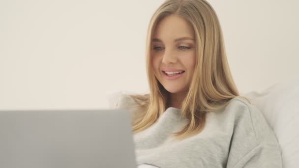 満足そうな笑顔若い女性あります探しています彼女のラップトップコンピュータで彼女の白いベッドで自宅で午前中 — ストック動画
