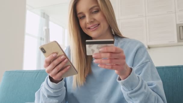幸せな若いですブロンドの女性あります使用しています彼女のスマートフォンながらホールドAカードで自宅のリビングルーム — ストック動画