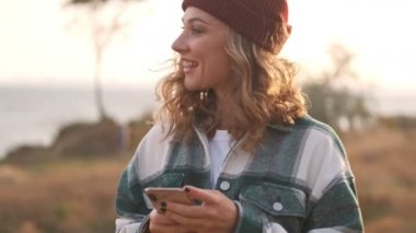 Şapka ve ekose gömlek giyen, akıllı telefon kullanan ve dışarı bakan mutlu sarışın kadın.