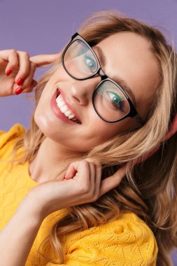 Gözlüklü, gülümseyen ve mor arka planda izole edilmiş kablosuz kulaklık kullanan neşeli bir kadın görüntüsü