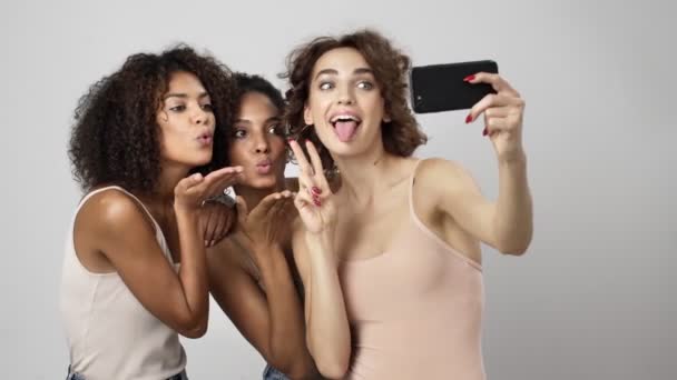 3人の美しい多民族の女性が白い背景に隔離されたスマートフォンで自撮り写真を撮っています — ストック動画