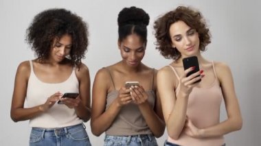 Üç hoş, çok ırklı kadın beyaz arka planda tek başına duran akıllı telefonlarını kullanıyor.