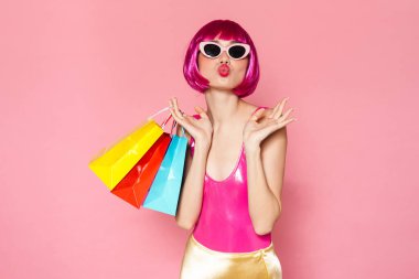 Renkli parlak kıyafetler giymiş, pembe arka planda izole edilmiş alışveriş çantaları taşıyan heyecanlı genç bir kadının portresi.