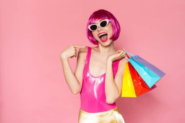 Renkli parlak kıyafetler giymiş, pembe arka planda izole edilmiş alışveriş çantaları taşıyan heyecanlı genç bir kadının portresi.