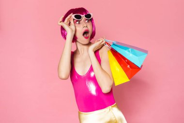 Pembe arka planda izole edilmiş alışveriş çantaları taşıyan renkli parlak kıyafetler giyen heyecanlı genç bir kadının portresi.