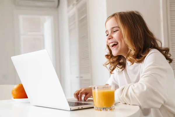 照片上 一个快乐可爱的女孩在笔记本电脑上打字 一边笑 一边在厨房里喝果汁 — 图库照片