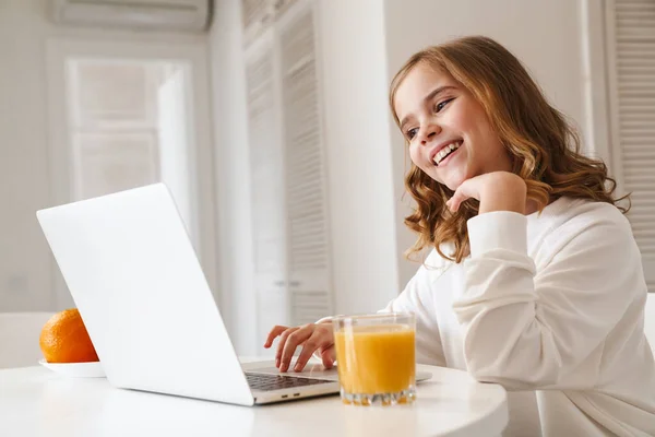 照片中快乐可爱的女孩在笔记本电脑上打字 在白色厨房喝果汁时微笑 — 图库照片