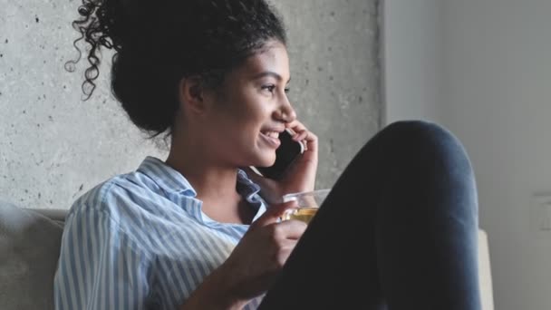 一位积极向上的年轻非洲妇女正坐在沙发上喝茶 一边用智能手机与人交谈 — 图库视频影像