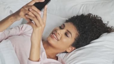 Gülümseyen genç bir Afrikalı kadın akıllı telefonunu evde yatağında kullanıyor.