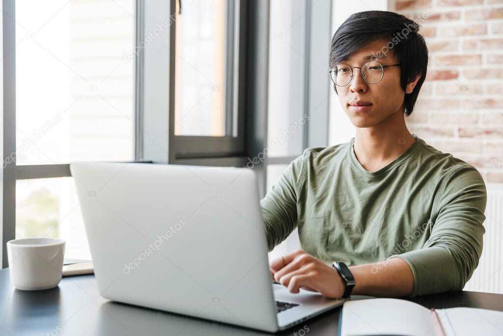 Image of handsome young asian man wearing eyeglasses using laptop while sitting at table in apartment
