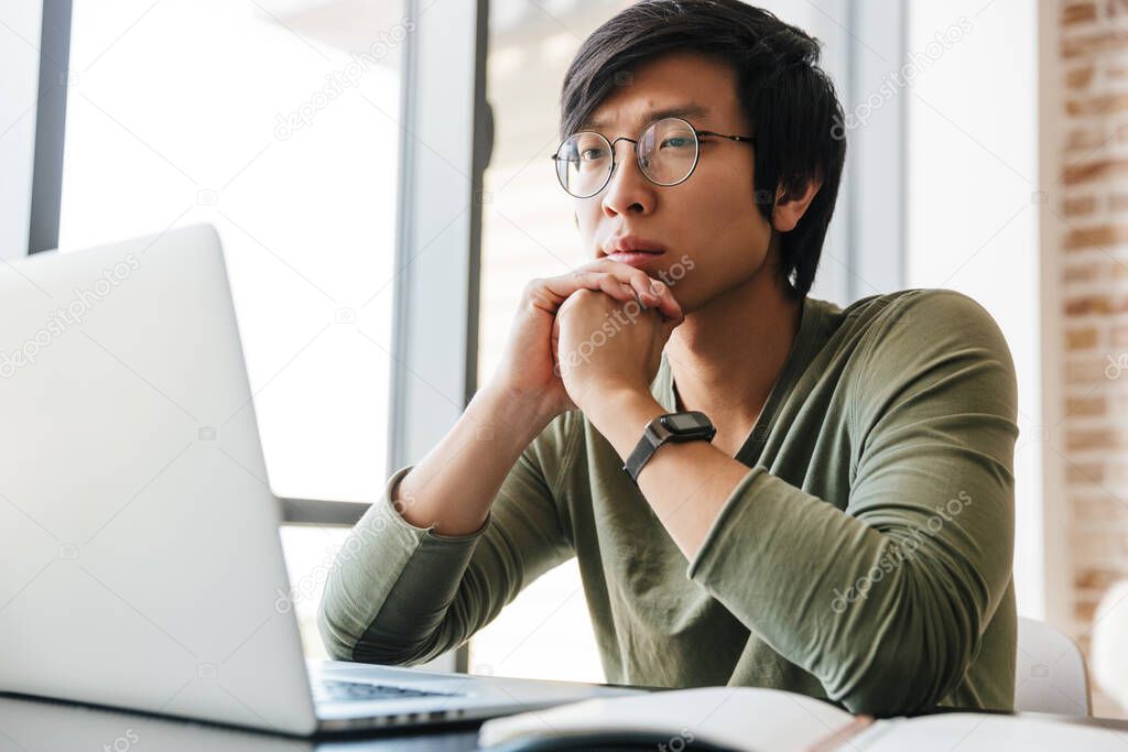 Image of handsome young asian man wearing eyeglasses using laptop while sitting at table in apartment