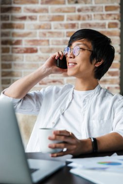 Gözlük takan, cep telefonu kullanan ve ofiste kahve içen yakışıklı bir Asyalı resmi.