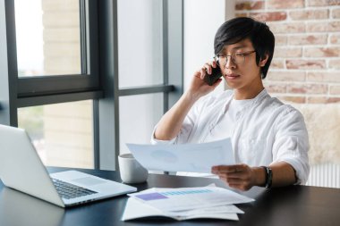 Gözlük takan, cep telefonu kullanan ve ofisinde evrak tutan yakışıklı Asyalı bir adam resmi.