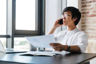 Gözlük takan, cep telefonu kullanan ve ofisinde evrak tutan yakışıklı Asyalı bir adam resmi.