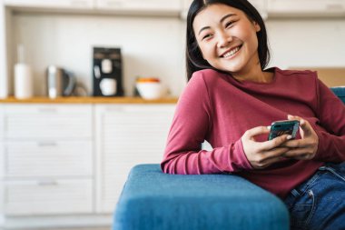 Gülümseyen Asyalı kadın evdeki koltukta otururken cep telefonu kullanıyor.