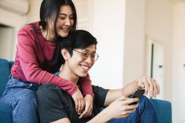 Evdeki koltukta oturan neşeli Asyalı çift cep telefonu kullanıyor.