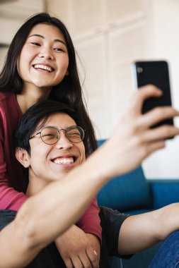 Neşeli Asyalı çiftin göz kırparken ve rahat bir odada otururken cep telefonunda selfie çekerken fotoğrafı.