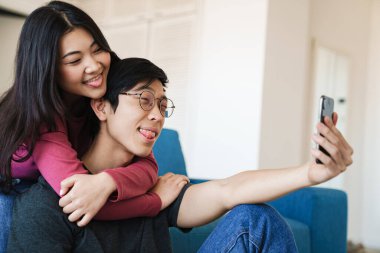 Komik Asyalı çiftin dillerini gösterdikleri ve odada otururken cep telefonlarından selfie çektikleri bir fotoğraf.