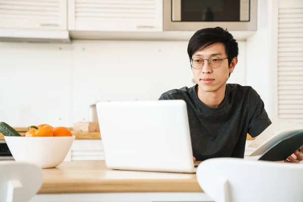 若い濃縮アジア人男性読書本の写真やノートパソコンを使用しながら テーブルの屋内に座って — ストック写真