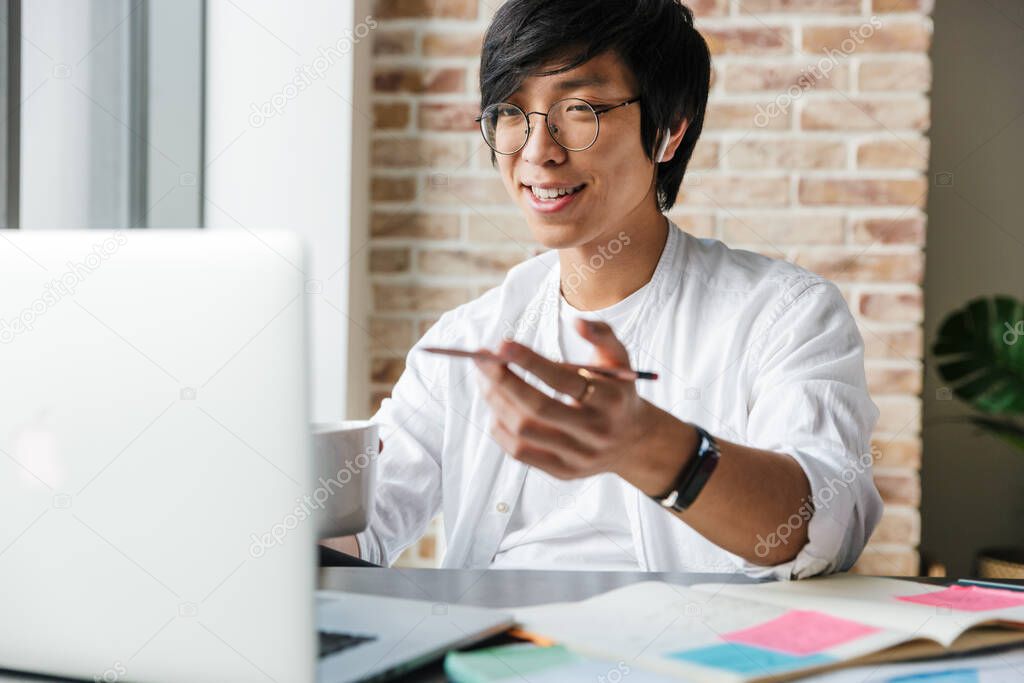 Image of handsome young asian man wearing earphones using laptop while sitting at table in office