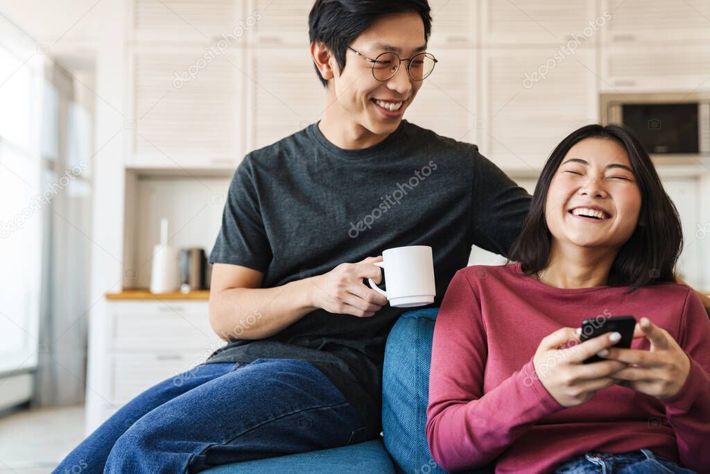 Cheerful young asian couple sitting on a couch at home, woman using mobile phone, man drinking coffee