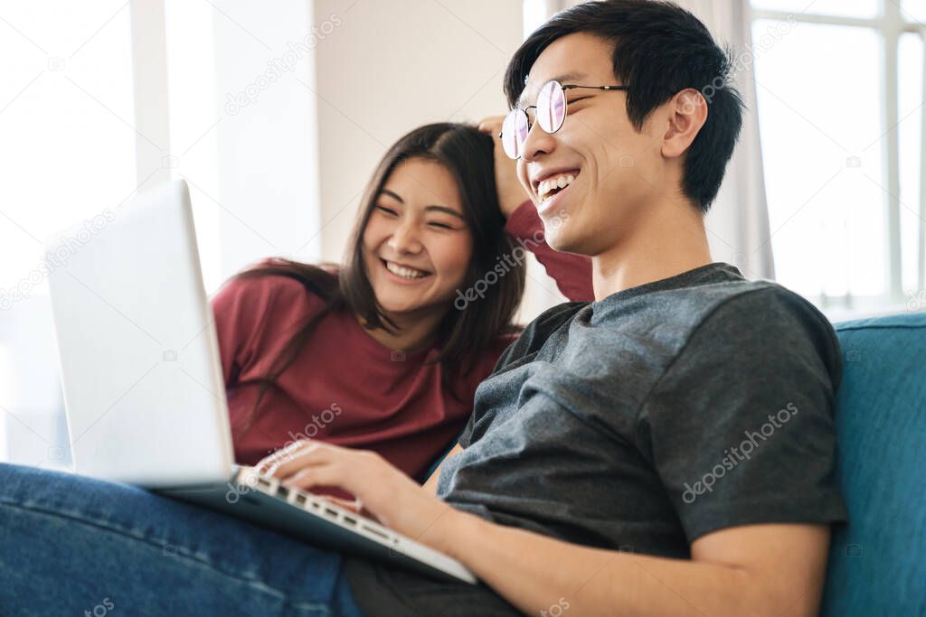 Cheerful young asian couple sitting on a couch at home, using laptop computer