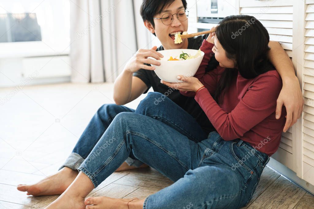 Cheerful young asian couple sitting on a floor in the kitchen, eating from a bowl