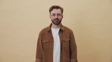 Tıraşsız, mutlu bir genç adam renkli alımlarını ve kredi kartını stüdyodaki bej duvar arkasında izole bir şekilde gösteriyor.