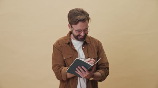 Uśmiechnięty Młody Nieogolony Mężczyzna Pisze Coś Swoim Copybooku Stojąc Samotnie — Wideo stockowe