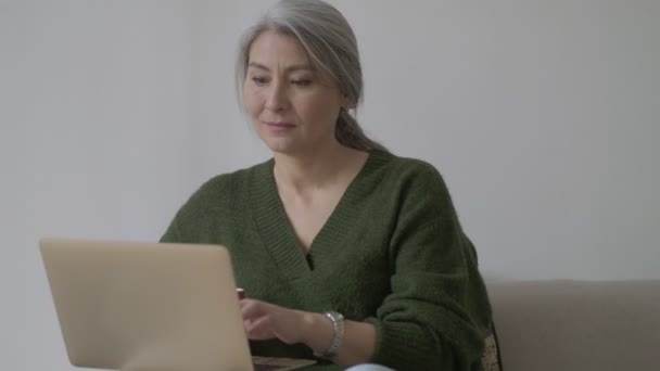 ソファの上にお茶を飲みながら屋内に座っている間 ラップトップコンピュータを使用して長い灰色の髪を持つ魅力的な成熟した女性を笑顔 — ストック動画