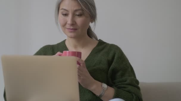 Uśmiechnięta Skoncentrowana Dojrzała Kobieta Długimi Siwymi Włosami Używająca Laptopa Pijąca — Wideo stockowe