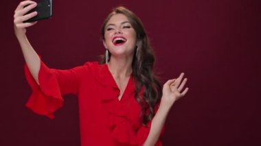 Kırmızı elbise giyen şık ve çekici genç bir kadın şarap rengi arka planda izole edilmiş akıllı telefonunu kullanarak selfie çekiyor.