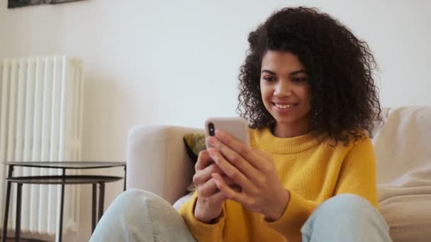 Sorrindo Africano Mulher Atraente Usando Smartphone Enquanto Sentado Chão Sala — Vídeo de Stock