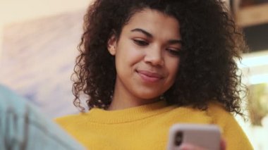 Akıllı telefon kullanan mutlu Afrikalı çekici bir kadın. Sonra da içeride sandalyede otururken kameraya bakıyor.