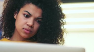 Ciddi, düşünceli, Afrikalı çekici bir kadın. İçeride sandalyede otururken dizüstü bilgisayar kullanıyor.