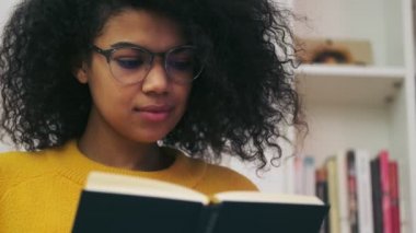 Kapalı alanda otururken gözlüklü, düşünceli Afrikalı çekici kadının kitap okuyuşuna yakından bakın.