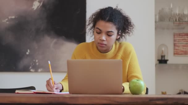 穏やかなアフリカの魅力的な女性ノートパソコンで何かを書き テーブルに座っている間にラップトップコンピュータを使用して屋内 — ストック動画