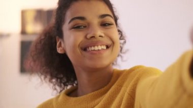 Sarı kazaklı neşeli Afrikalı kadının oturma odasında poz verişini ve selfie çekişini yakından izleyin.