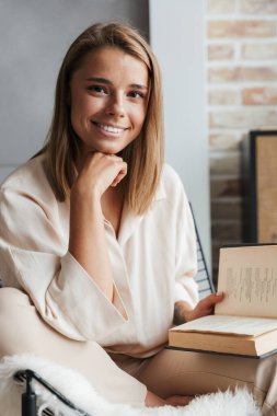 Oturma odasındaki koltukta otururken şık pijamalarıyla kitap okuyan, gülümseyen genç bir kadın resmi.